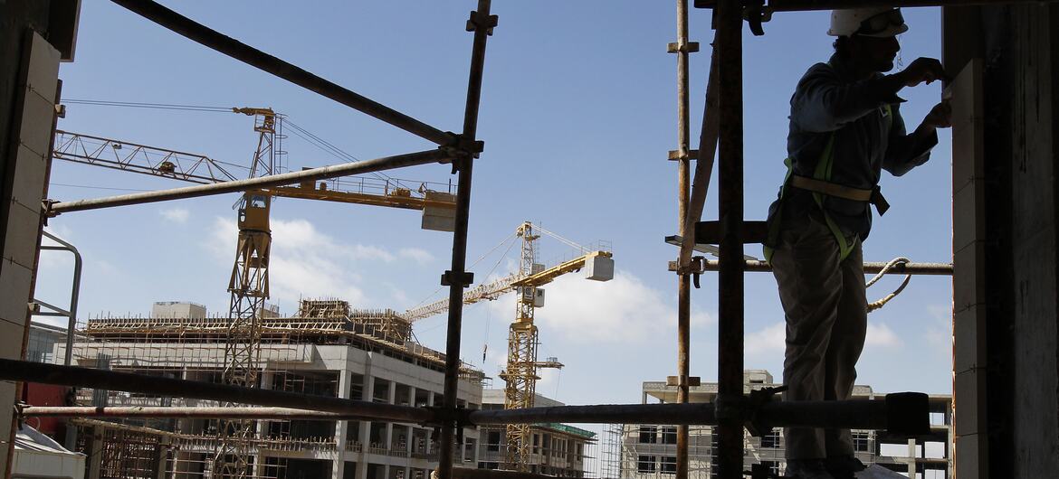一名头戴安全帽、身系安全带的建筑工人站在建筑工地的脚手架上，背景中有起重机和正在施工的建筑。