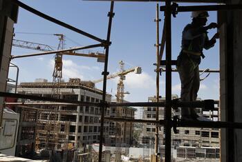 A construction worker wearing a hard hat and safety harness stands on scaffolding at a construction site with cranes and a building under construction in the background.
