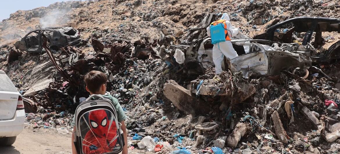 A young boy with a Spiderman backpack walks past a person in a white hazmat suit spraying disinfectant on a pile of waste and destroyed cars in Gaza.