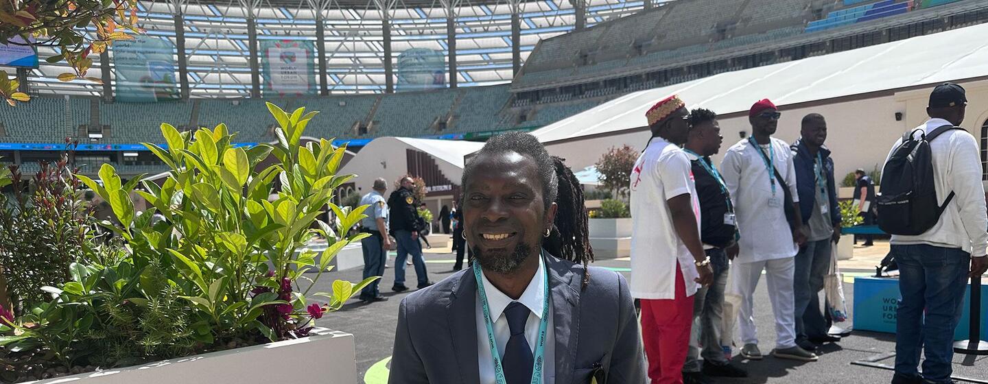 A smiling Black man in a suit and lanyard sits on a white bench at an outdoor event in a stadium.