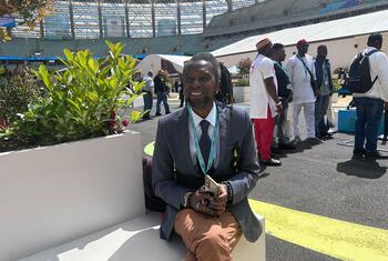 A smiling Black man in a suit and lanyard sits on a white bench at an outdoor event in a stadium.