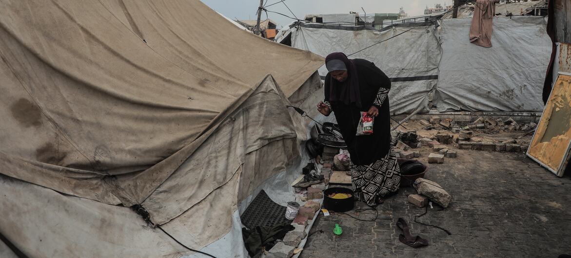 A woman standing outside a weathered tent prepares food. She stands in an area surrounded by makeshift tents and improvised shelters at a settlement in Gaza City.