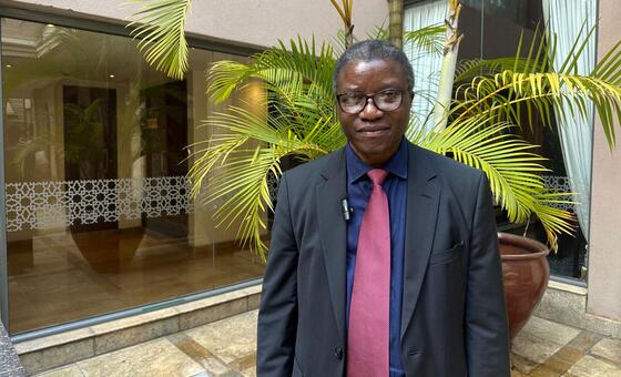 Mshauri maalum wa Umoja wa Mataifa wa kuzuia mauaji ya kimbari, Chaloka Beyani, amesimama mbele ya jengo la kioo lenye mimea ya mitende, akiwa amevaa suti na shati la waridi.