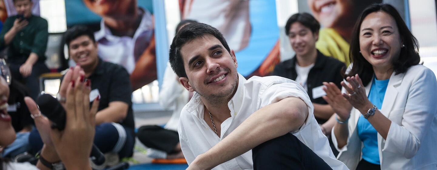 Un joven sonriente con una camisa blanca se sienta frente a un grupo de personas aplaudiendo, en un evento de diálogo juvenil celebrado en la oficina de UNICEF en Tailandia en febrero de 2024.