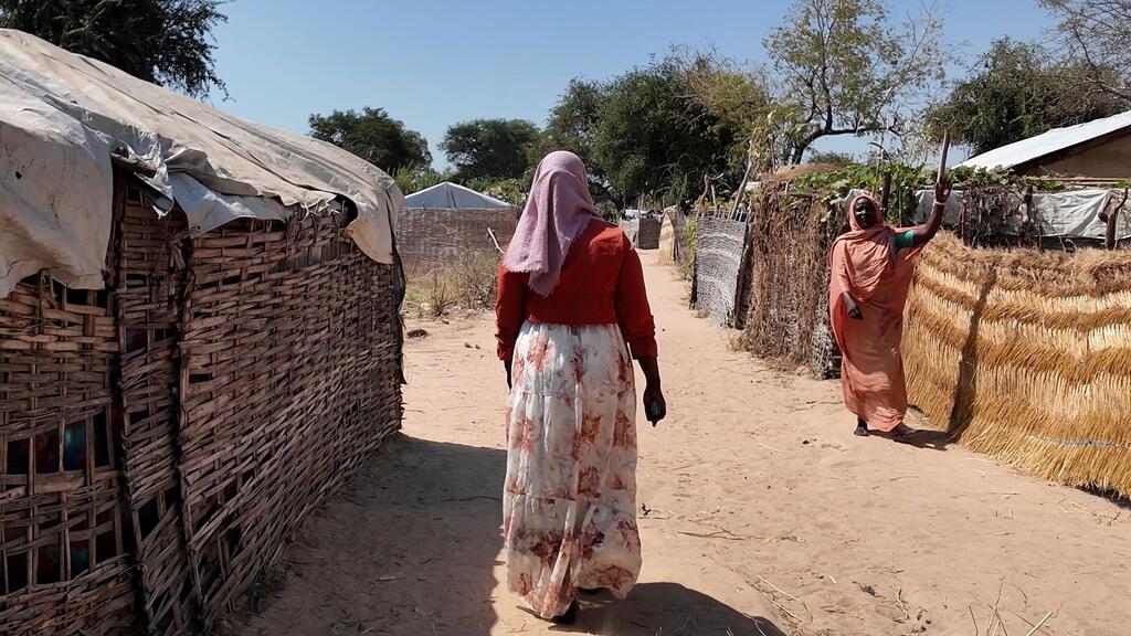 A woman walks through a refugee camp in the Central African Republic, with another woman waving in the background. The scene highlights displacement due to conflict in Sudan.