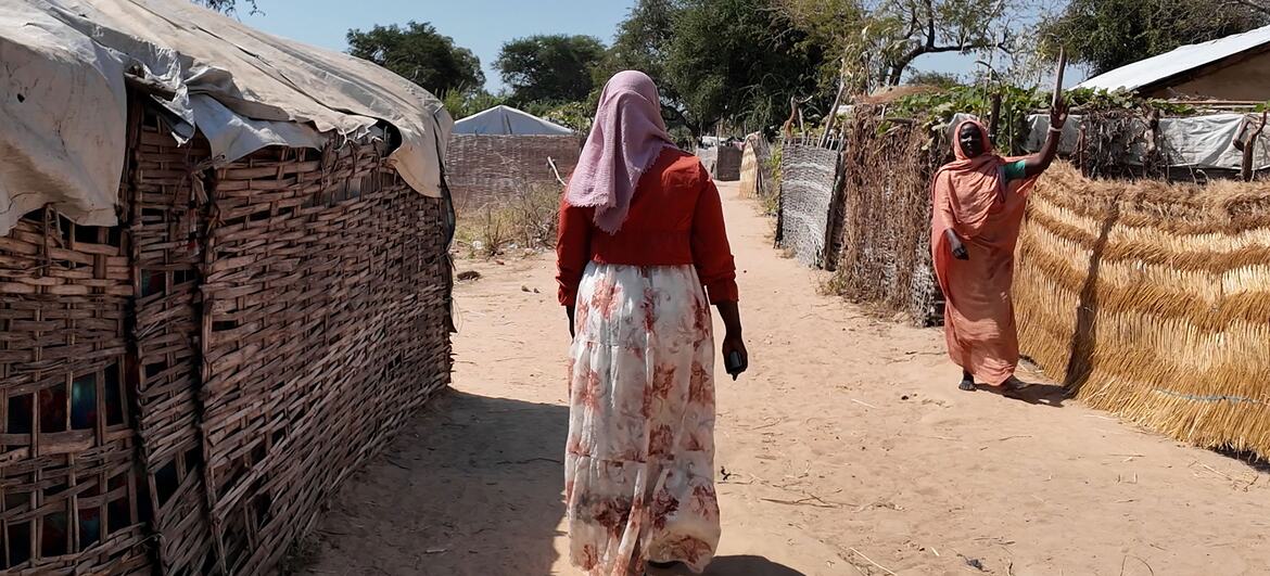 A woman walks through a refugee camp in the Central African Republic, with another woman waving in the background. The scene highlights displacement due to conflict in Sudan.