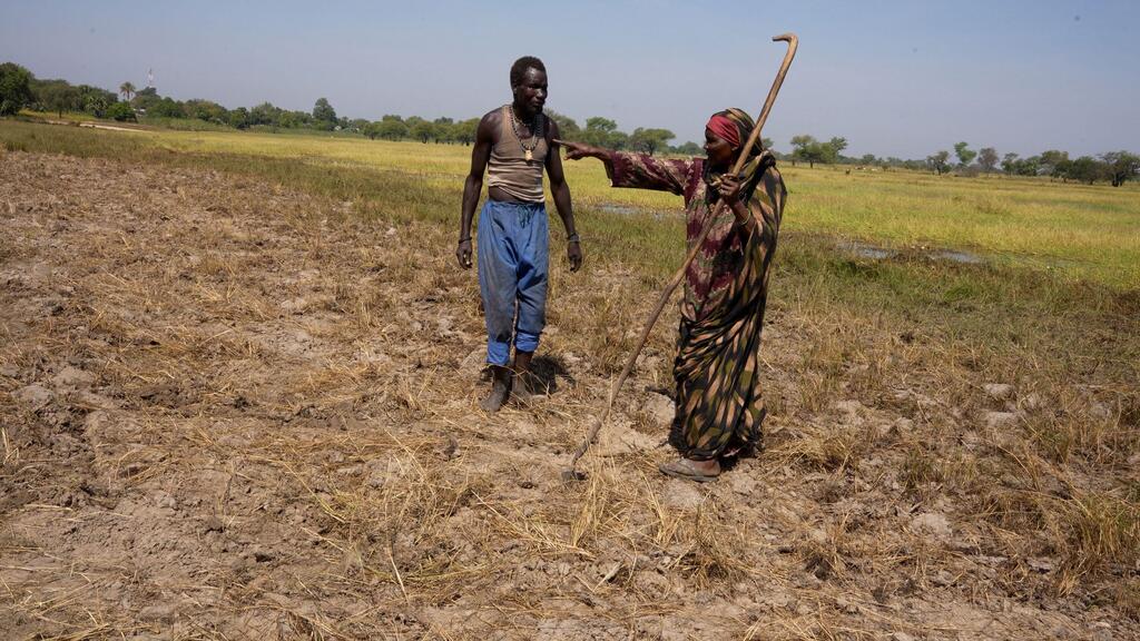 A Sudanese refugee woman teaches a man how to work the field in the Central African Republic, demonstrating survival techniques using a traditional farming tool.