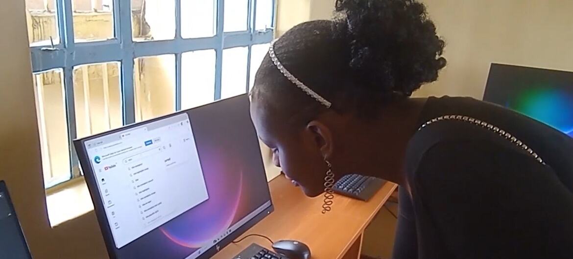 Uma jovem com cabelo trenzado e um acessório de flores rosa está sentada em um computador, enquanto uma mulher adulta se inclina para ajudá-la. O cenário parece ser um centro de aprendizagem de TIC em Kajiado, Quênia, com uma janela no fundo.