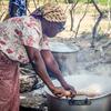 Joana Maria João Chongo, de 71 anos, uma mulher afetada pelas inundações em Chókwè, Moçambique, cozinha farinha de milho em uma grande panela sobre uma fogueira aberta no centro de alojamento de Chiaquelane, destacando sua resiliência apesar dos repetidos deslocamentos por inundações.