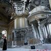 Un homme vêtu d'une robe noire et d'un chapeau orange se tient à l'intérieur d'une église endommagée et partiellement détruite à Odessa, en Ukraine. L'intérieur orné montre des signes de dégâts de guerre avec des dômes brisés, des poutres exposées et des débris.