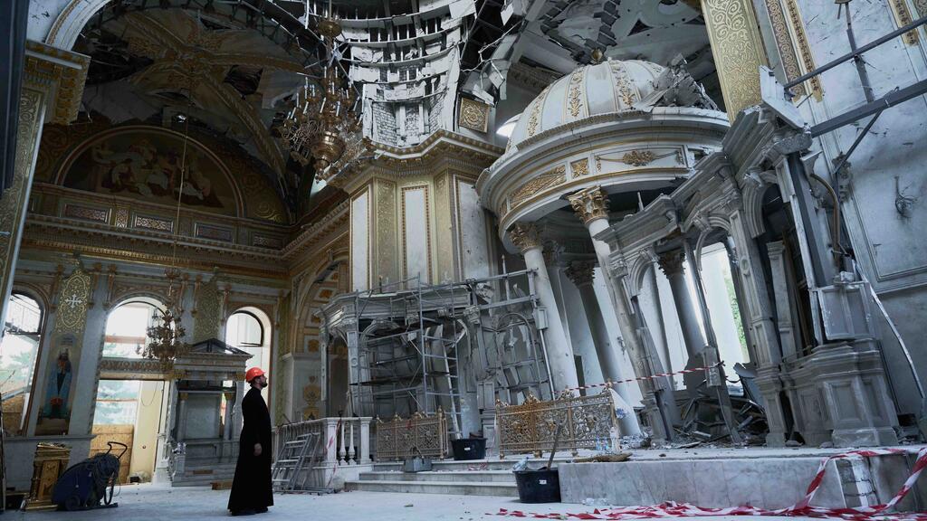 Un hombre de túnica negra y sombrero duro naranja se encuentra dentro de una iglesia dañada y parcialmente destruida en Odessa, Ucrania. El interior adornado muestra signos de daños de guerra con cúpulas rotas, vigas expuestas y escombros.