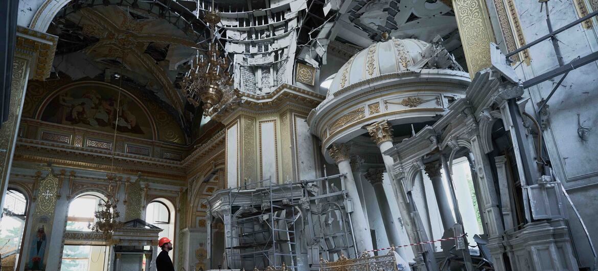 Un homme vêtu d'une robe noire et d'un chapeau orange se tient à l'intérieur d'une église endommagée et partiellement détruite à Odessa, en Ukraine. L'intérieur orné montre des signes de dégâts de guerre avec des dômes brisés, des poutres exposées et des débris.