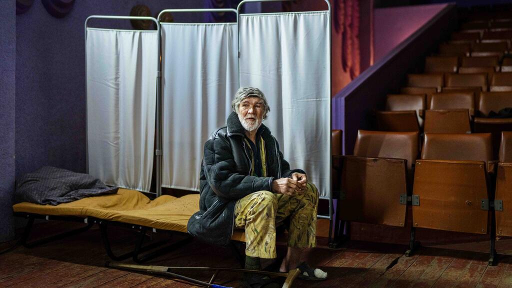 An elderly Ukrainian man sits on a cot in a theater-turned-shelter, surrounded by rows of empty seats and privacy screens, highlighting the humanitarian crisis caused by the ongoing war.