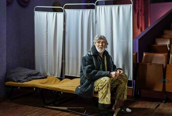 Un homme ukrainien âgé est assis sur un lit dans un refuge transformé en théâtre, entouré de rangées de sièges vides et d'écrans de protection de la vie privée, soulignant la crise humanitaire causée par la guerre en cours.