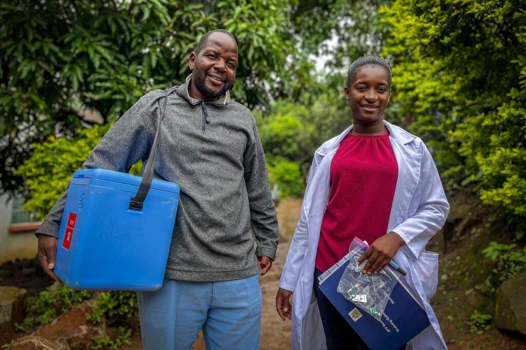 Wafanyakazi wawili wa afya nchini Malawi, mmoja akichukua chombo cha bluu cha kukomesha chanjo na mwingine akichukua vifaa vya matibabu, wanatabasamu mbele ya kamera.