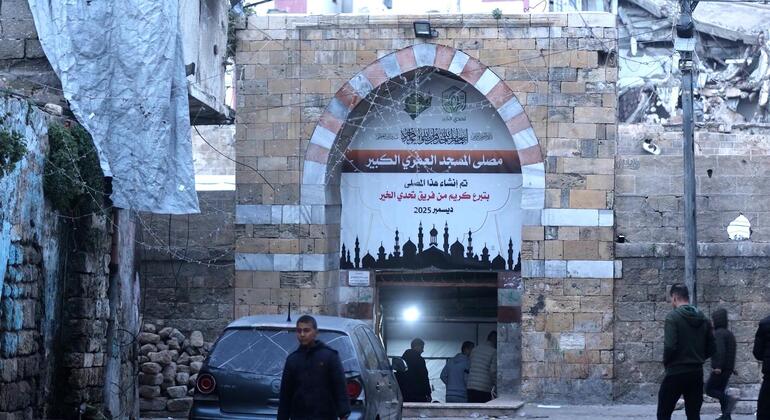 منظر من مدخل المسجد الأقصى في غزة، تظهر أضرار حربية كبيرة في المباني المحيطة. علقة لافتة كبيرة مع نص عربي فوق القوس الحجري، ويجتاز عدد قليل من الرجال سيارة متضررة.