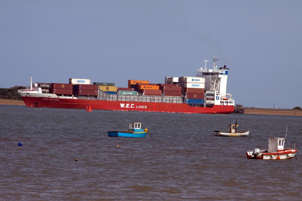 تبحر سفينة حاويات كبيرة من شركة W.E.C. LINES باللونين الأحمر والأبيض، محملةً بحاويات شحن ملونة، على الماء، يرافقها ثلاثة قوارب أصغر.