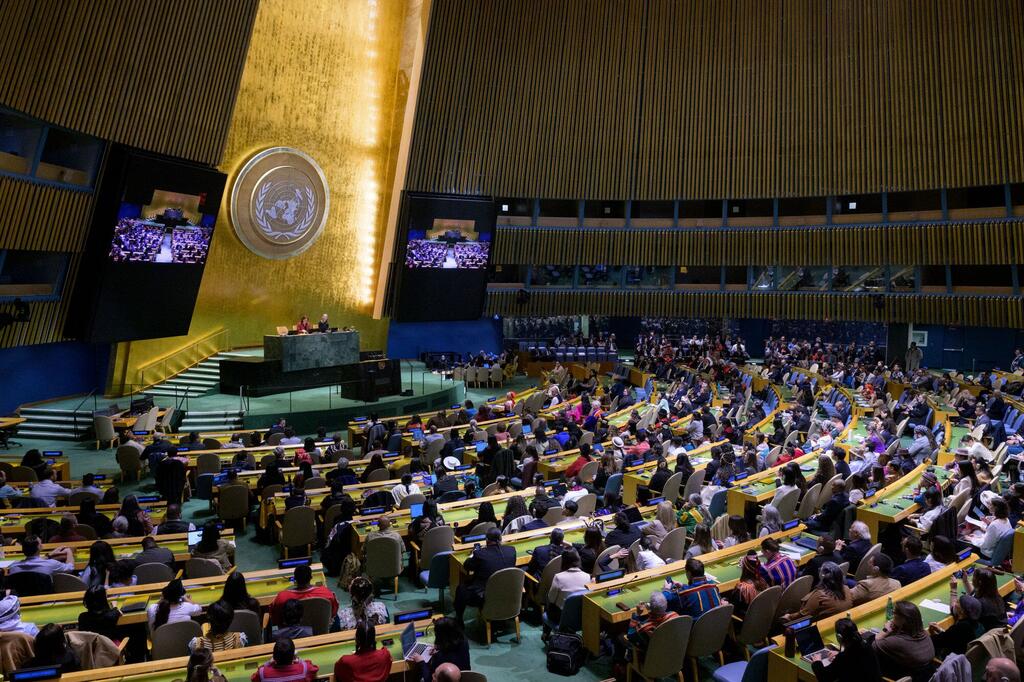 联合国大会大厅全景，正值联合国原住民问题常设论坛（UNPFII）开幕之际。大厅内座无虚席，与会者坐在弧形桌前，面向悬挂着联合国大徽标的中央讲台。