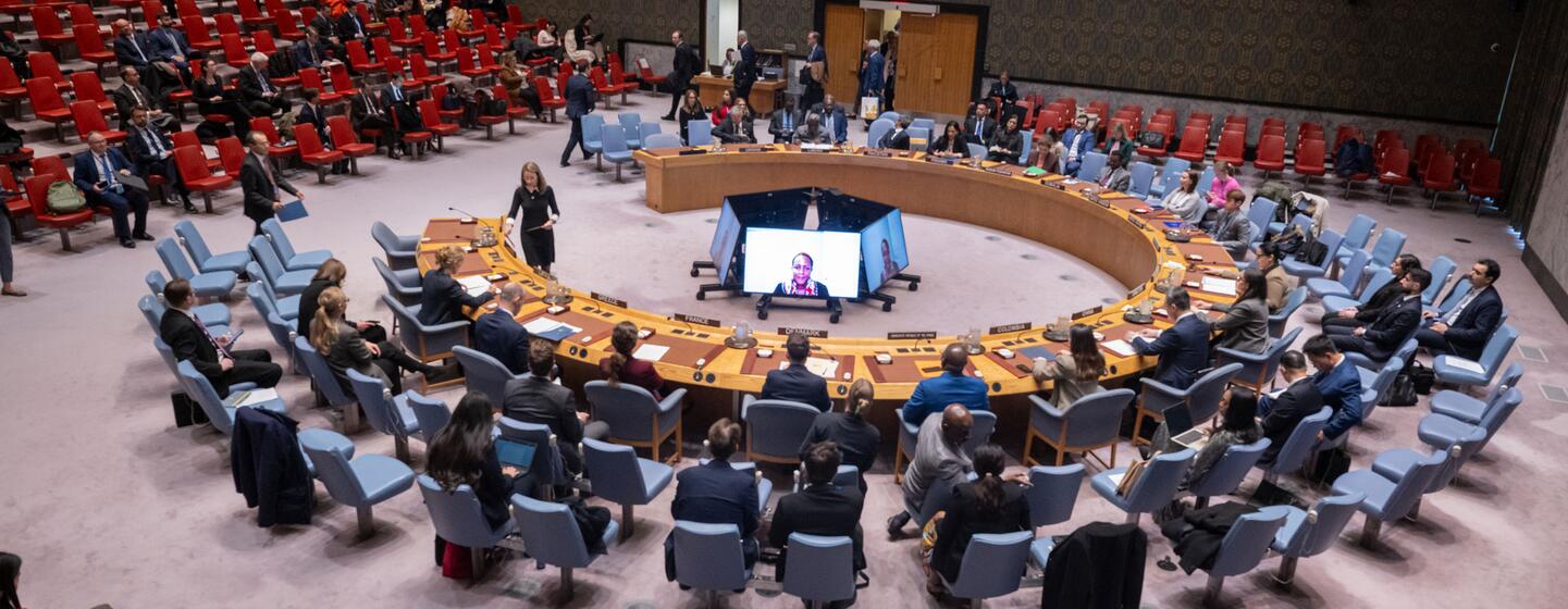 Une vue large et en contre-plongée de la salle du Conseil de sécurité des Nations unies lors d'une réunion sur le maintien de la paix et de la sécurité en Ukraine. Les délégués sont assis autour de la table en fer à cheval emblématique, avec un flux de vidéoconférence visible sur un écran au centre.