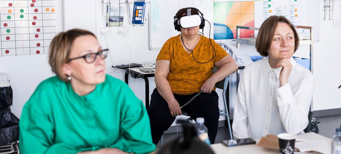 A woman in an orange shirt wearing a VR headset sits at a table while two other women observe. The setting appears to be a meeting or presentation about healthcare technology.