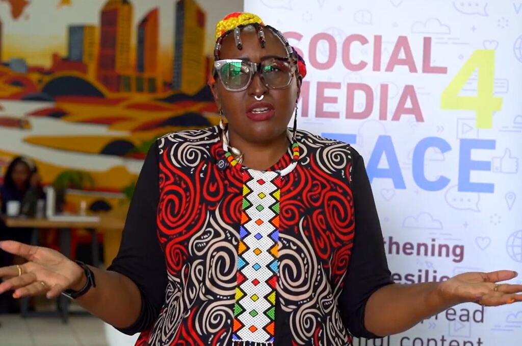 Mwanamke mweusi aliyevaa miwani na koti la rangi za kitamaduni la Kiafrika anazungumza katika tukio la UNESCO kuhusu deepfakes na mitandao ya kijamii nchini Kenya.