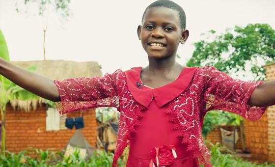 Msichana kijana Mwafrika aliyevaa sare nyekundu ya shule anatabasamu akiwa na mikono iliyonyooshwa katika mazingira ya vijijini nchini Tanzania.