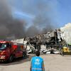 Un trabajador del ACNUR observa un edificio de almacén destruido con humo que sale de él, con un camión de bomberos rojo y una cargadora amarilla cerca.