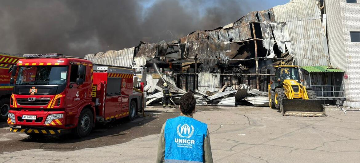 Um funcionário do ACNUR observa um armazém destruído com fumaça saindo dele, com um caminhão de bombeiros vermelho e uma escavadeira amarela nas proximidades.