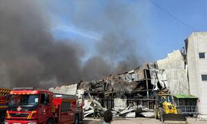 یو این ایچ سی آر کا ایک ملازم ایک تباہ شدہ گودام کی عمارت کو دیکھ رہا ہے جس سے دھواں اڑ رہا ہے، قریب میں ایک سرخ فائر ٹرک اور ایک پیلا لوڈر ہے۔