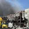 A damaged UNHCR warehouse in Dnipro, Ukraine, with black smoke rising from the burnt structure and a yellow excavator in the foreground.