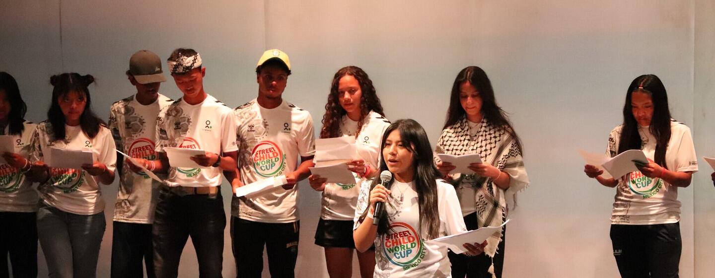 Un grupo de jóvenes está en un escenario durante un evento en México: una chica en primer plano habla en un micrófono mientras sostiene unos papeles, y otras personas están detrás de ella, también con papeles.