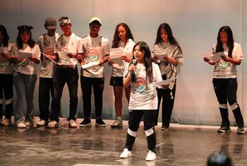 Un grupo de jóvenes está en un escenario durante un evento en México: una chica en primer plano habla en un micrófono mientras sostiene unos papeles, y otras personas están detrás de ella, también con papeles.