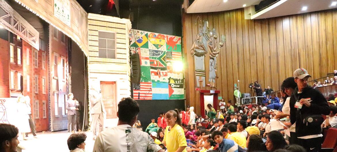 Niños y adultos observan una función escénica en un auditorio. Un niño en primer plano lleva una camisa blanca con "Pakistán" en la espalda.
