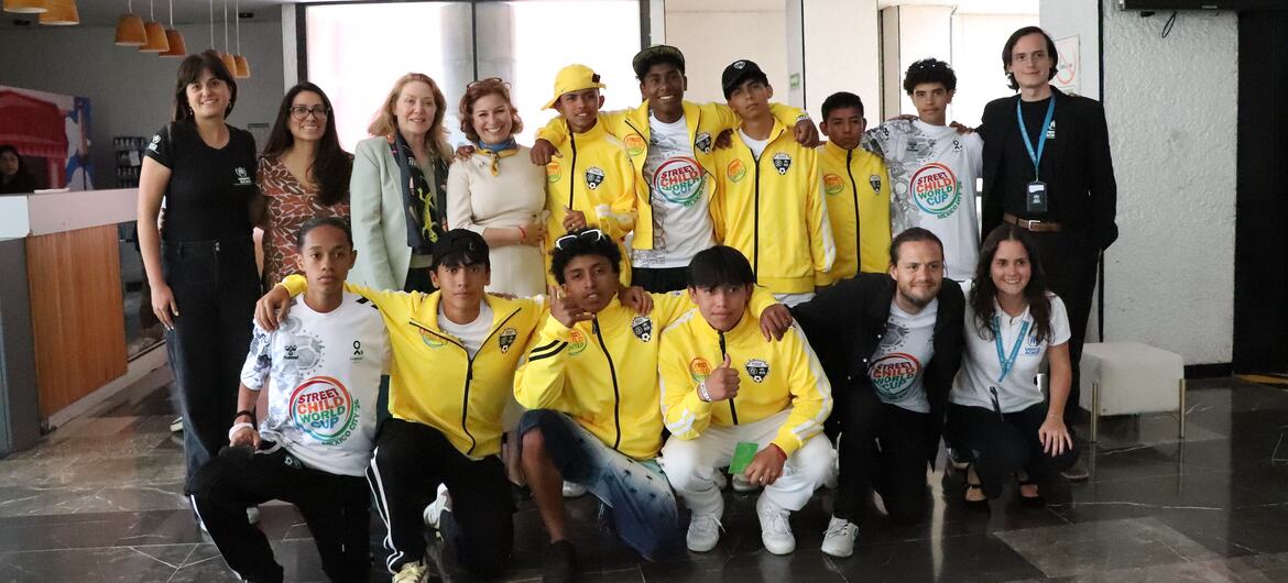 Un grupo de jóvenes jugadores de fútbol con chaquetas amarillas y camisetas blancas posa para una foto con funcionarios de UNICEF en la oficina de UNICEF México.