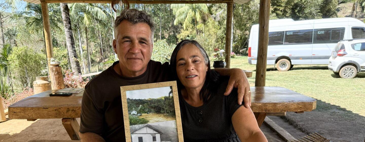 Um casal, Henrique e Tania, sentados em uma mesa de madeira ao ar livre, segurando uma foto emoldurada de uma casa e alguns papéis, cercados por palmeiras e veículos estacionados.