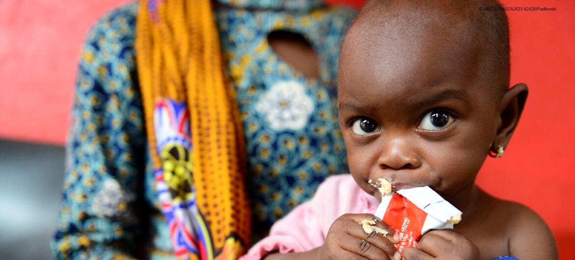 Uma criança africana sendo segurada por uma mulher enquanto comia de um pacote de ajuda alimentar vermelho e branco.