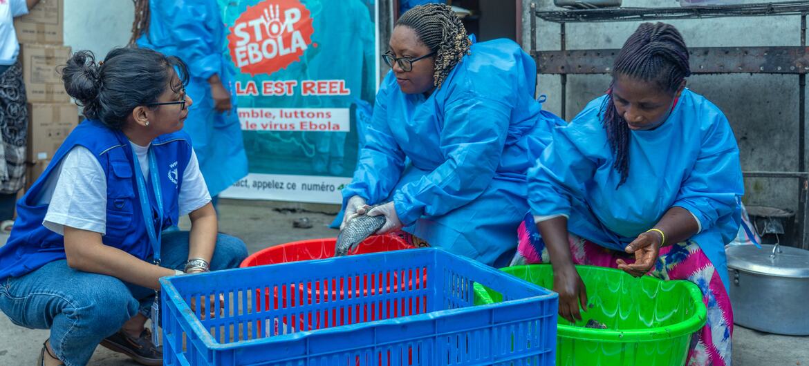 Three women in blue protective suits prepare food from green and red plastic basins. A fourth woman in a blue vest and jeans crouches nearby, observing them.