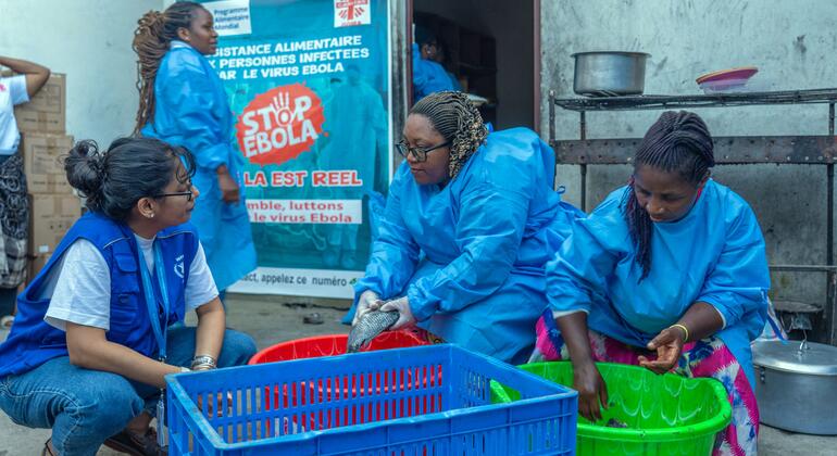 Tres mujeres con trajes protectores azules preparan comida en recipientes de plástico verdes y rojos. Una cuarta mujer, con un chaleco azul y pantalones vaqueros, se agacha cerca, observándolas.