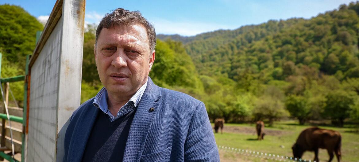 Мужчина в синем костюме стоит перед полем, на заднем плане видны бизоны.