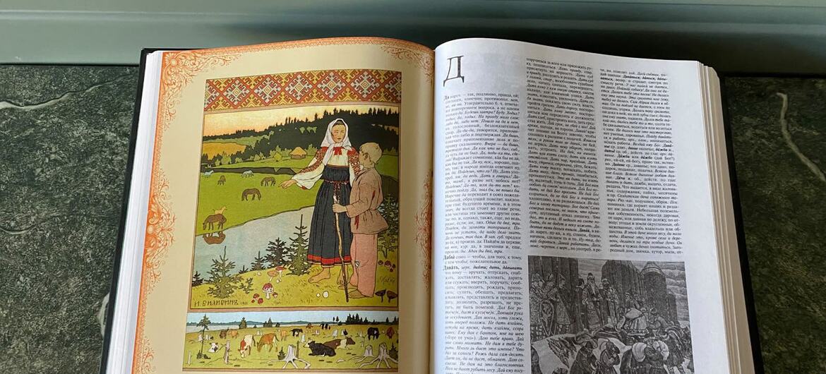 На темной поверхности лежит открытая книга с русским текстом и красочными иллюстрациями, изображающими русских крестьян и сельские пейзажи.