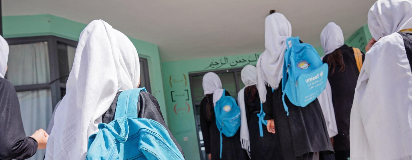 A group of female students wearing hijabs and carrying UNICEF-branded blue backpacks walk towards a school building.