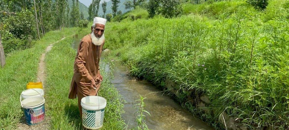 روایتی لباس میں ملبوس ایک بزرگ پاکستان کے ایک دیہی، پہاڑی علاقے میں ندی سے بالٹی بھر رہے ہیں۔