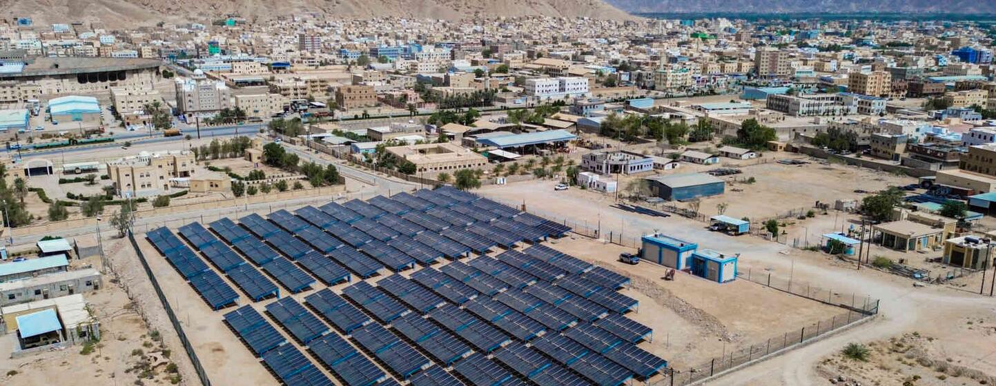 Aerial view of a solar panel array in a desert landscape near a city in Yemen, with a large mountain in the background.