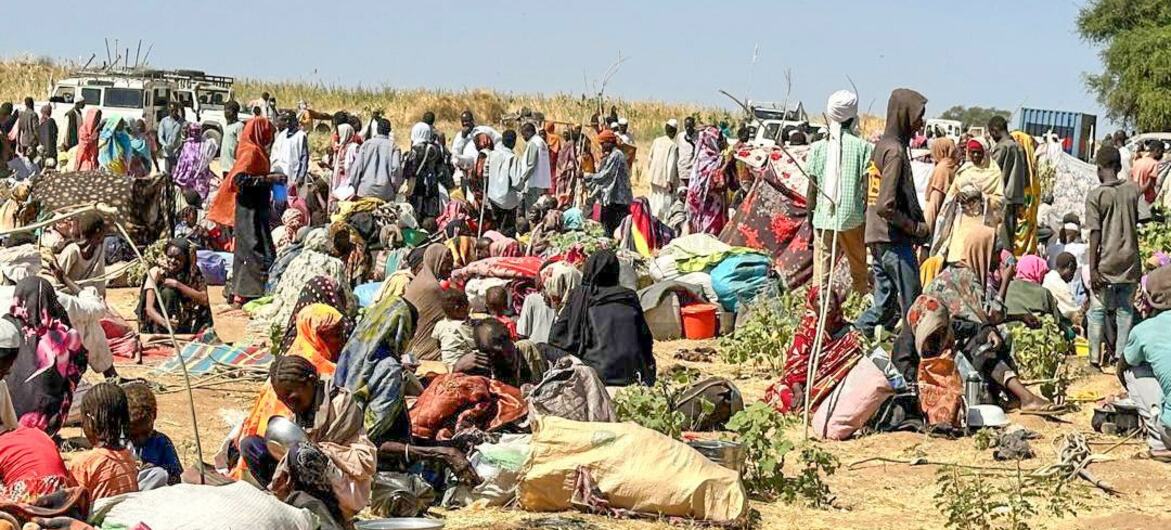Des personnes déplacées se rassemblent dans un camp de réfugiés au Soudan, avec des travailleurs humanitaires et des véhicules des ONU visibles en arrière-plan.
