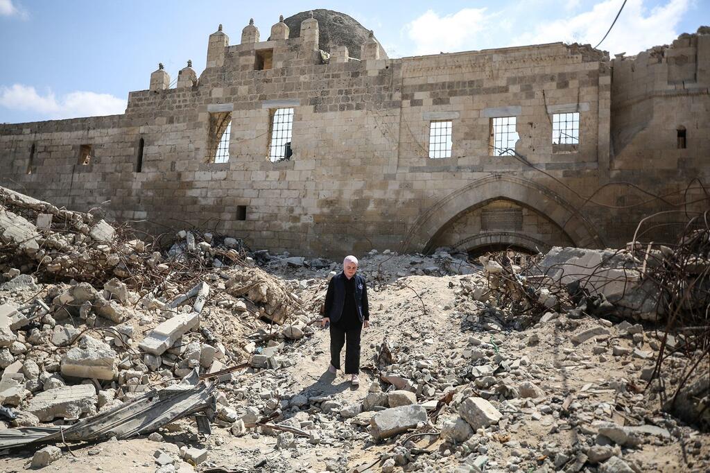 Um homem de jaqueta e calças escuras está parado em um caminho de entulho em frente a um grande edifício de pedra danificado, com uma cúpula e uma porta em arco, em Gaza.