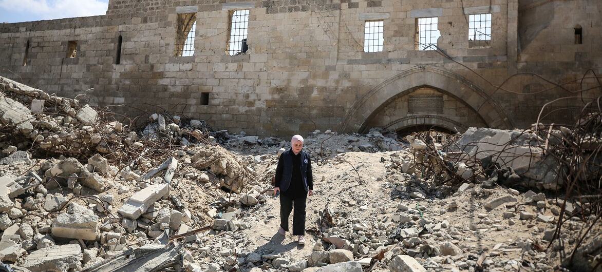 À Gaza, un homme vêtu d'une veste et d'un pantalon sombres se tient sur un chemin de décombres devant un grand bâtiment en pierre endommagé, surmonté d'un dôme et d'une porte cintrée.