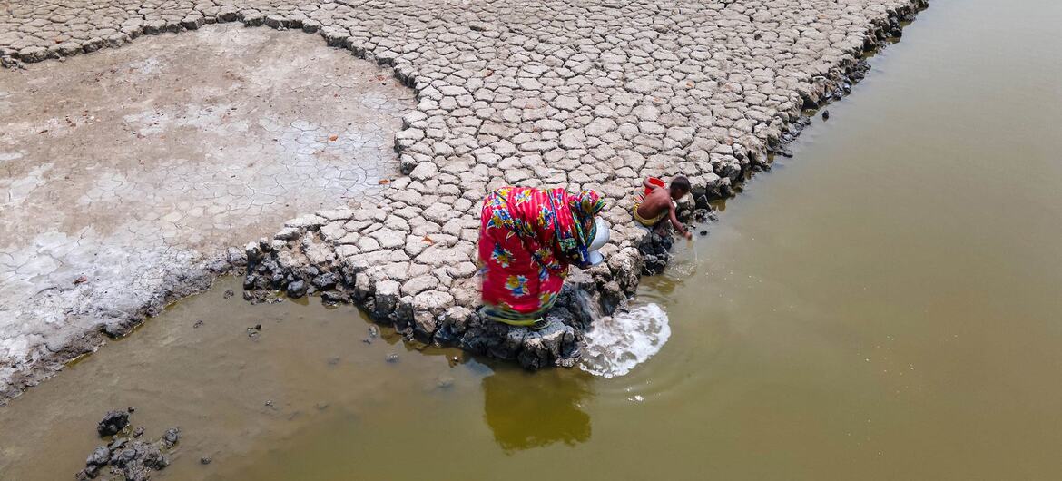 Uma mulher e uma criança buscam água em uma poça turva na orla de uma vasta paisagem árida e rachada em Bangladesh.