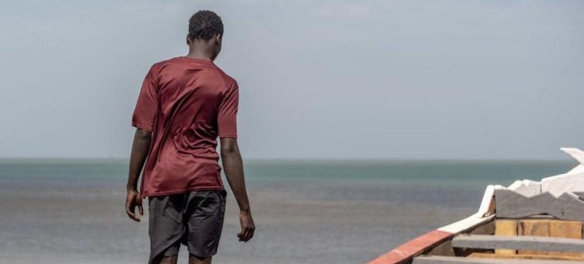 Un hombre se aleja de la cámara caminando por el borde de una barca de madera en una playa de arena con el océano de fondo.