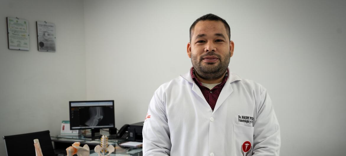 Un médico hispano, vestido con una bata blanca, se encuentra en un consultorio médico, con modelos anatómicos de una rodilla y una columna vertebral sobre un escritorio detrás de él.