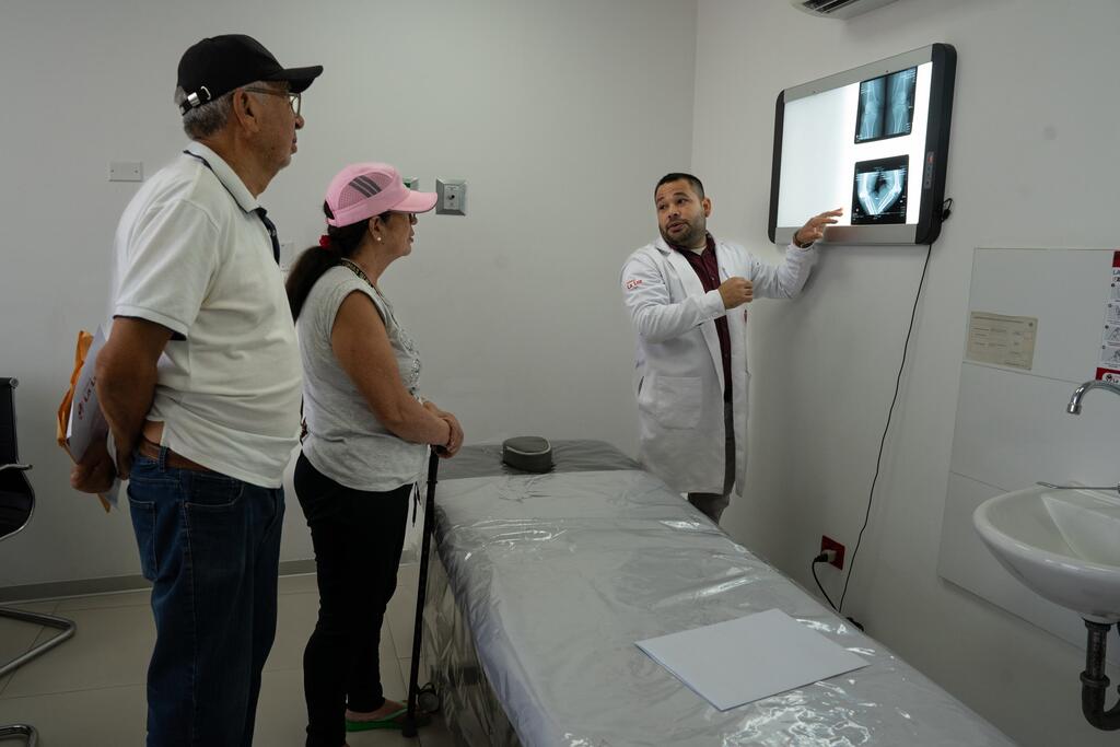 Un médico con bata blanca señala las radiografías en una pantalla mientras se las explica a un hombre y una mujer mayores en una sala de consulta.